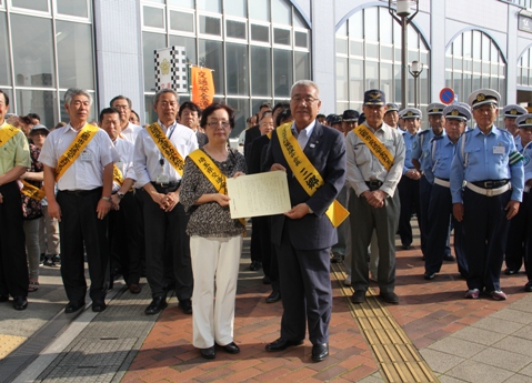 警察官や関係者の方々が並んで立っている前で、黄色のタスキを肩にかけた田村さんと市長が一緒に交通安全啓発知事メッセージの用紙を持っている写真