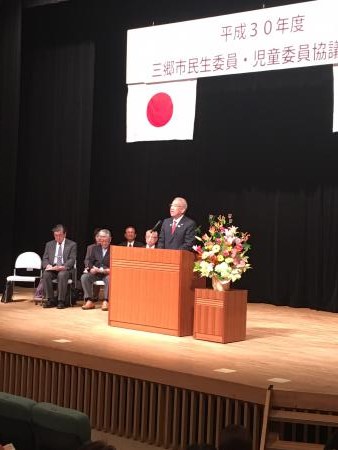 三郷市民生委員・児童委員協議会総会の吊り看板の設置された舞台上で、演壇の前に立って話をしている市長の写真