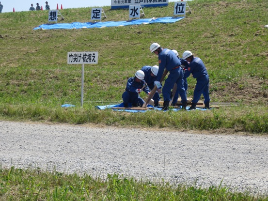 芝生の生えた土手に「竹尖げ・杭拵え」と書かれた看板が設置され、活動服とヘルメットを着用した関係者たちがビニールシートの上で作業をしている写真