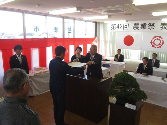 「第42回農業祭表彰式」と書かれた横断幕と国旗・三郷市旗が吊り下げられている会場で、立派な盆栽が置かれた花台と並ぶ演台に立ち市長賞を手渡す三郷市長と向かい合って立ち両手で受け取る受賞者、市長の近くに立つ介添え人の女性の写真