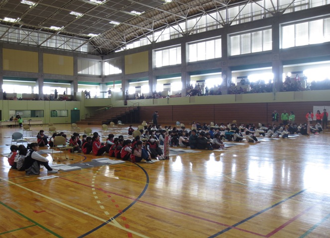総合体育館の床に敷かれたグレーのシートの横に体育座りで整列している赤いゼッケンを着用した子どもたちとゼッケンを着用していない子どもたちの写真