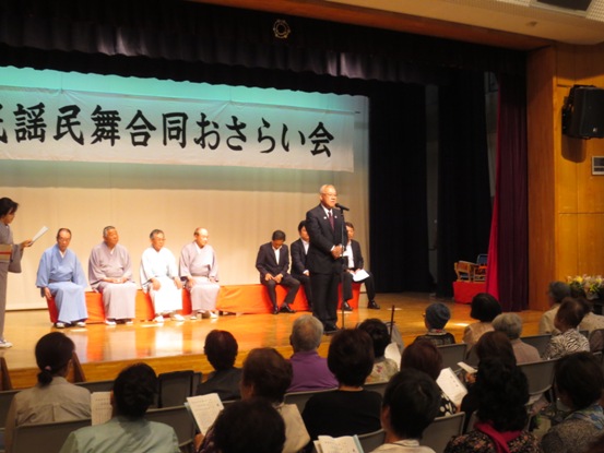 民謡民舞合同おさらい会の横断幕が設置された舞台上に、衣装を着た4名の男性やスーツ姿の男性たちが後ろに座っており、市長がマイク台の前に立って参加者たちに話をしている様子の写真