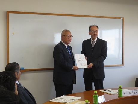壁に設置されたホワイトボードの前に立つ三郷市長と野中会長がともに諮問書を持って記念撮影をしている写真