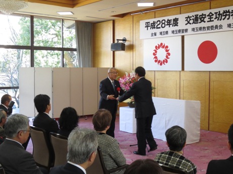「平成28年度交通安全功労者等表彰式」の横断幕と埼玉県の旗、国旗が掲げられた壁の前で、埼玉県知事から表彰を受ける市長と、その様子を見ている椅子に着席した参加者たちの写真