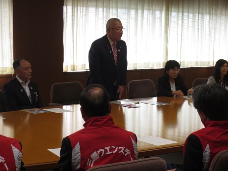席を立ち挨拶の言葉を述べる三郷市長と、資料が置かれた席に座り話を聞く関係者と白字で「三郷ウエンズデー」と書かれた赤いベストを着用した三郷ウェンズデーロータリークラブのメンバーの写真