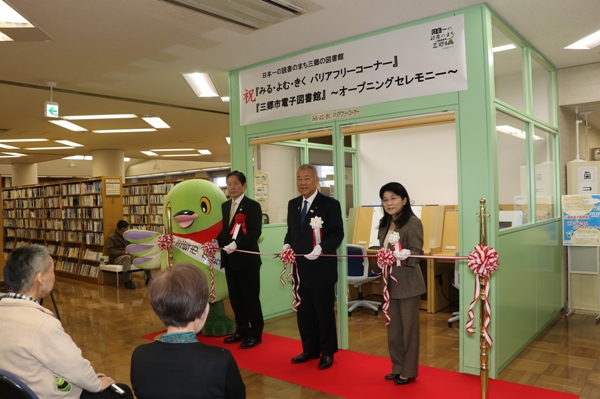 「祝『みる・よむ・きくバリアフリーコーナー』『三郷市電子図書館』オープニングセレモニー」と書かれたポスターが貼りつけられている、窓のついた薄緑色の壁で仕切られたバリアフリーコーナーの前で、レッドカーペットの上に立ちテープカットセレモニーを行う三郷市長と関係者、白いタスキをかけたかいちゃんの写真