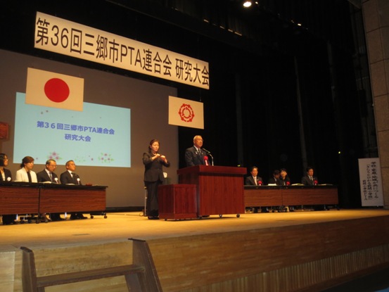 「第36回三郷市PTA連合会研究大会」の吊り看板が設置されている舞台上で、両側に関係者たちが着席しているなか、中央の演台に立って話をしている市長の写真