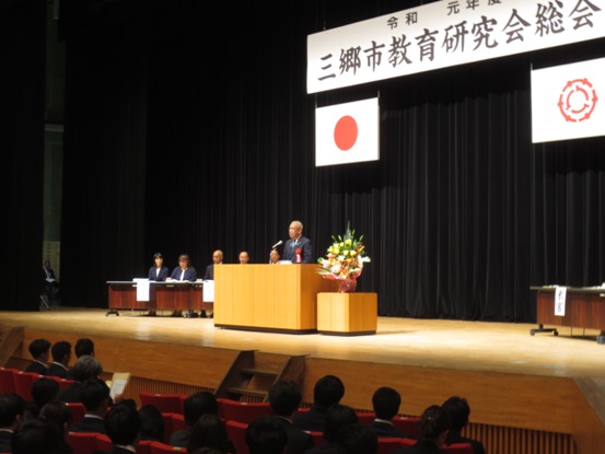 「三郷市教育研究会総会」と書かれた吊り看板が設置された舞台上で、演壇に立って挨拶をする市長と、客席に座っている多くの関係者たちの写真
