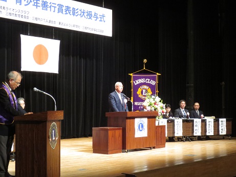舞台上に左手前から、司会者台、演台、ライオンズクラブの旗、来賓席が並んでおり、中央の演台で市長が挨拶をしている様子の写真