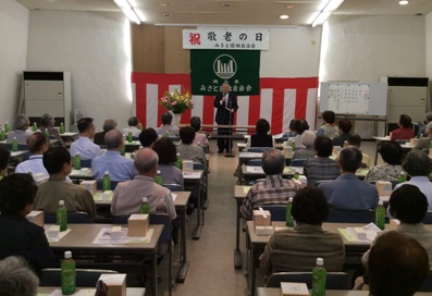 「祝敬老の日」と書かれ紅白幕が貼られた三郷団地中央集会所で挨拶を行っている市長の話を着席して聞いている参加者の写真