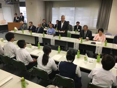 室内に設置された長テーブルの席に中学生と関係者が向かい合って座り、関係者側中央の席から立ち上がり挨拶を行っている市長の写真