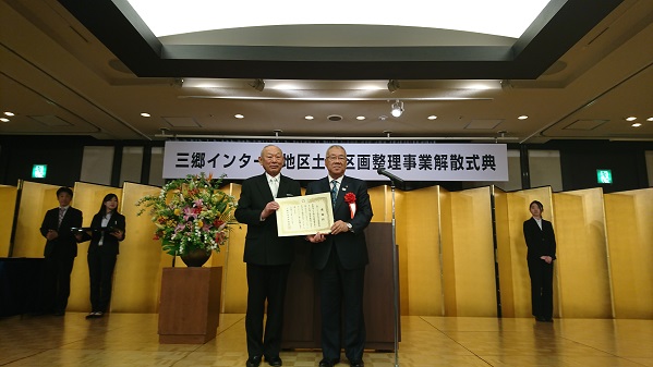「三郷インターA地区土地区画整理事業解散式典」と書かれた横断幕が天井から吊り下げられ、金色のパーテーションや赤・ピンク・黄色・緑・白など色とりどりの花がアレンジされた大きな花壺が置かれた会場で、三郷市長が組合の関係者と感謝状を片方ずつ持ち合い記念撮影をしている写真