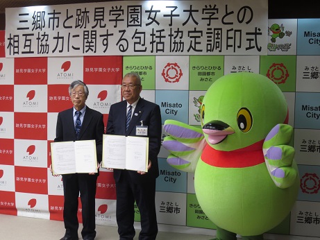 「三郷市と跡見学園女子大学との相互協力に関する包括協定調印式」と書かれたポスターが貼られた三郷市と跡見学園女子大学のボードの前で、三郷市長と跡見学園女子大学の関係者が協定書を手に持ち、かいちゃんとともに記念撮影をしている写真