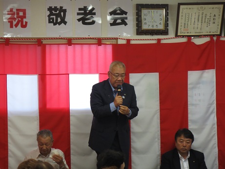一文字ずつ「祝 敬老会」と書かれ紅白幕が貼られた室内で、立ち上がった市長が右手にマイクを持ち挨拶を行っている写真