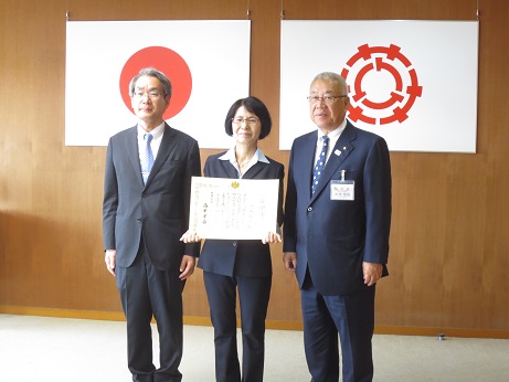 国旗と三郷市旗が吊り下げられた壁の前で、三郷市長と感謝状を手に持った會田さん(中央)、関東管区行政評価局の男性(左)が記念撮影をしている写真
