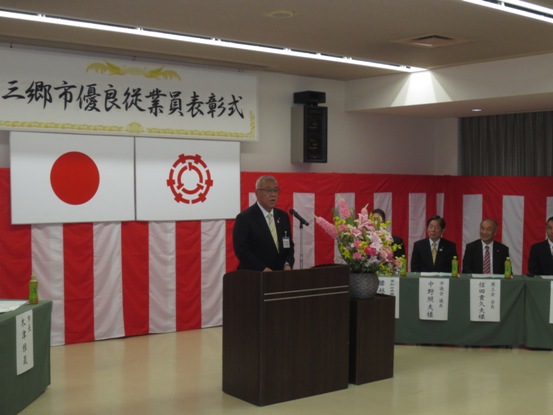 「三郷市優良従業員表彰式」と書かれたボードと国旗・三郷市旗が吊り下げられている壁の前で、ピンク・黄色・白の豪華な花々で彩られた花台と並ぶ演台に立ち式辞を述べる三郷市長と、紅白幕近くに配置された緑のカバーが掛けられ席札が貼りつけられている席に座り話を聞く関係者の写真