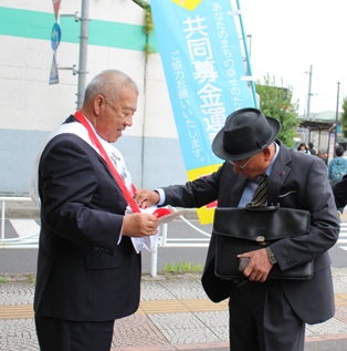 赤い羽根共同募金ののぼり旗が設置された前で、スーツ姿の男性が市長が持っている募金箱に募金を行っている様子の写真