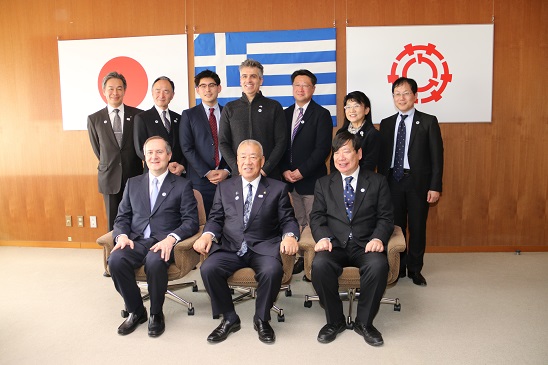 日本国旗・ギリシャ国旗・三郷市旗が吊り下げられている壁の前で、三郷市長と駐日ギリシャ共和国大使館、日本大学、日本ギリシャ協会の関係者が並んで記念撮影をしている写真