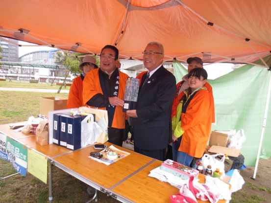 バザーテントの下で、三郷市長がオレンジ色の法被を着て農産品を販売している福島県広野町の関係者とともに記念撮影をしている写真