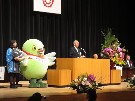 三郷市の旗が掲げられた舞台上の、大きな花が飾られた演台に立ち客席に向かって式辞を述べる市長と、その隣に立つかいちゃんとつぶちゃん、青色の法被を着た女性を写した写真