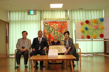 オレンジ色の着物が掛けられ、ホワイトボードに「祝100歳おめでとう」と書かれた模造紙が貼られている室内で、車いすに座って額縁に入った賞状を持った東ヶ崎きくいさんと市長、関係者の男性と女性が横一列に並んで座っている記念写真