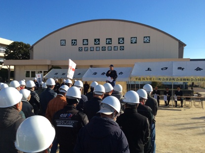 テントが並んだ朝礼台の上に市長が立ち話をしているのを向かい合って立っているヘルメットを被った参加者が聞いている後ろ姿の写真