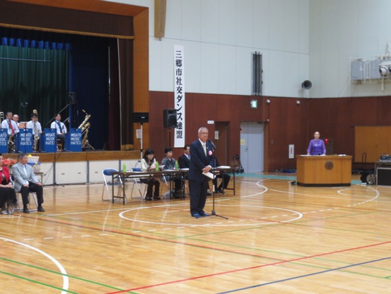 壁に「三郷市社交ダンス連盟」の紙が下がっている体育館で、後方でオーケストラのかたが舞台上に座っており、舞台下のマイク台の前に立って市長が話をしている写真