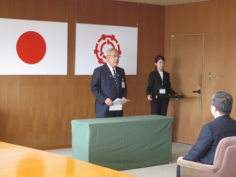 国旗と三郷市旗が吊り下げられている壁の前で、緑のカバーが掛けられた台の後ろに立ち感謝の言葉を述べる三郷市長と後方で賞状盆を持つ介添人の写真