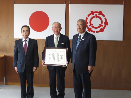 国旗と三郷市旗が吊り下げられている壁の前で、感謝状を手に持った三郷市文化協会の男性が三郷市長と関係者の男性とともに記念撮影をしている写真
