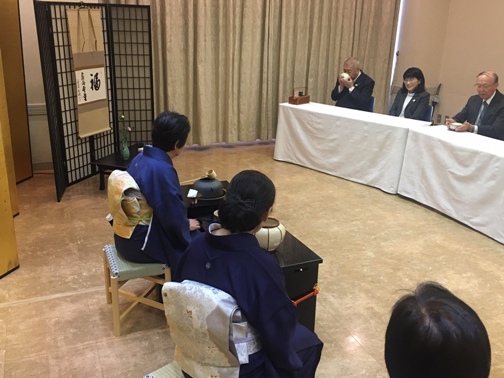 椅子に座る着物姿の女性たちが見守る中、向かいに設置された白いクロスの引かれたテーブル席に座った市長がお茶をいただいている写真