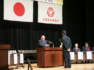 右側に胸章リボンをつけた関係者が座り、中央の演壇を挟んで男性と向かい合って立っている市長が表彰状を読み上げている写真