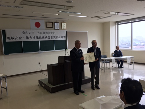 「令和元年 吉川警察署管内地域安全・暴力排除推進大会表彰伝達式」と書かれた横幕が貼られた室内の窓際の席に男性が座り、中央の演壇の前で市長と三郷鳰鳥ライオンズクラブの男性が一緒に感謝状を持っている記念写真