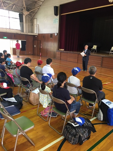 参加者たちが体育館に設置されたパイプ椅子に座り、正面に立って話をしている市長の方を見ている写真