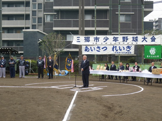 フェンスに「三郷市少年野球大会」の横断幕が張ってあるグラウンドで、バッターボックスに設置されたマイク台の前に立った市長が挨拶をしている写真