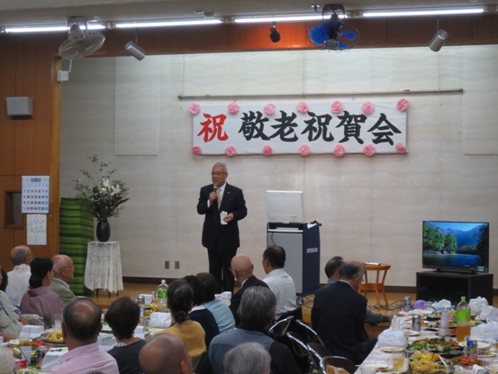 「祝 敬老祝賀会」の吊り看板が設置された会場で、多くの参加者が見守る中、市長がマイクを手に話をしている写真