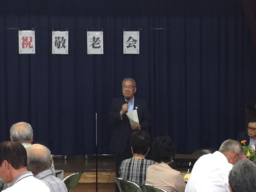 一文字ずつ「祝敬老会」と書かれた文字が貼られた会場の席に座っている参加者の前で、右手にマイクを持ち挨拶を行っている市長の写真