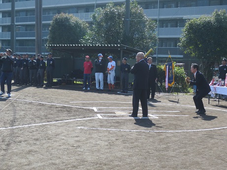 野球バットを持ちバッターボックスに立っている三郷市長と右手にグローブをはめ構える姿勢を取っている男性、始球式の様子を見守る子どもたちの写真