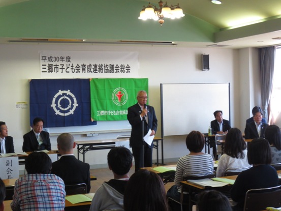 「平成30年度三郷市子ども会育成連絡協議会総会」と書かれた紙が貼ってある部屋で、等間隔に置かれた長テーブルに資料を置いた参加者たちが着席しているなか、市長が話をしている写真