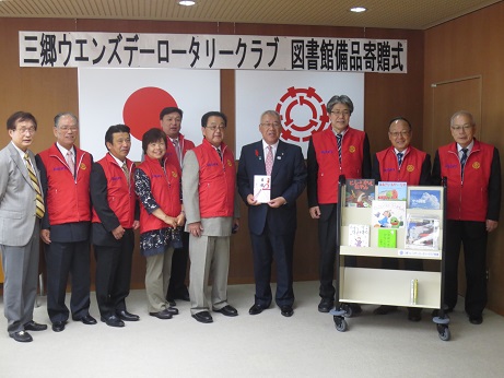 「三郷ウエンズデーロータリークラブ図書館備品寄贈式」と書かれた横断幕と国旗・三郷市旗が吊り下げられている壁の前で、祝儀袋を手に持った三郷市長と、おそろいの赤いベストを着用した三郷ウェンズデーロータリークラブのメンバーたちが、絵本が置かれたディスプレイラックとともに並んで立っている記念写真