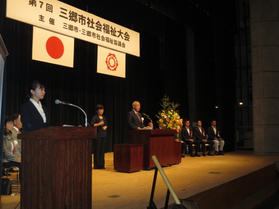 「第7回三郷市社会福祉大会」の吊り看板が設置されている薄暗い舞台上で、演壇に立って話をしている市長の写真