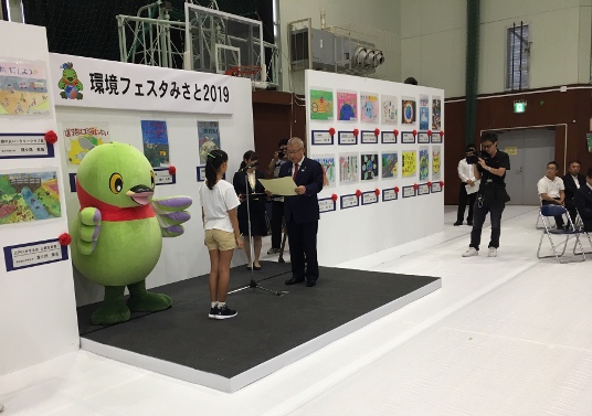 「環境フェスタみさと2019」と書かれた横幕が貼られた小さなステージ上でかいちゃんが見守る中、マイクスタンドを挟んで左側に女の子が立ち、右側に市長が立って表彰状を読んでいる写真