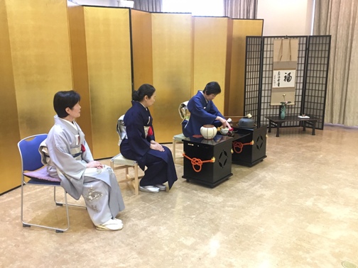 金屏風が設置された会場で、着物を着た3名の女性が椅子に座っており、右側に座る女性がお茶をたてている写真