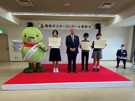 環境ポスターコンクールの表彰を受けた3名の子ども達と一緒に、赤い絨毯の上で記念撮影をする木津市長とかいちゃんの写真