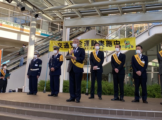 ららぽーと新三郷内ステージ上で、7名程の関係者と交通安全運動実施中の横断幕前で、マイクを持ち挨拶をする黄色いたすきをかけた木津市長の写真