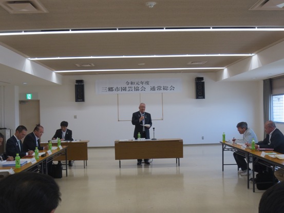 「令和元年度三郷市園芸協会通常総会」の紙が貼られた部屋で、関係者が四方から向かい合わせに着席しているなか、正面中央に立って挨拶をしている市長の写真