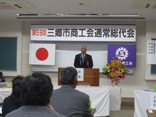 「第59回三郷市商工会通常総代会」の吊り看板が設置された部屋で、関係者が椅子に着席しているなか、演壇に立つ市長が挨拶をしている写真