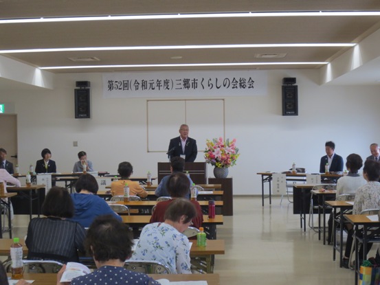壁に「第52回 令和元年度三郷市くらしの会総会」と書かれた紙が貼ってある部屋で、等間隔に並べられたテーブルに2名ずつの関係者が椅子に着席しているなか、正面の演壇に立って挨拶をしている市長の写真