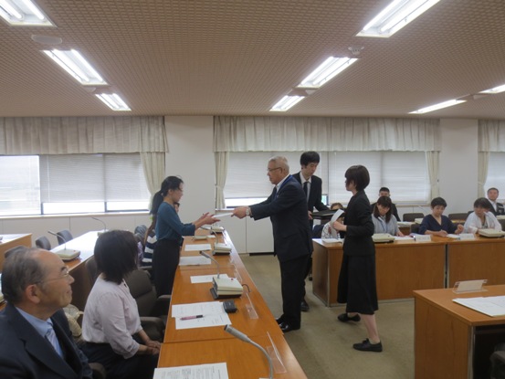 長机が並んでいる部屋で、椅子に着席している関係者たちと、一名の女性が市長から委嘱書の交付を受けている写真