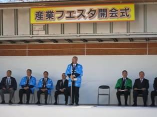「産業フェスタ開会式」と書かれた横断幕の下に設置されたマイクスタンドの前で挨拶を行っている市長の写真
