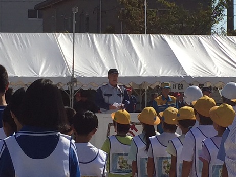 屋外に設置された白いテント前の演台に立ち、開会式で訓示する、災害時用の制服を着用した三郷市長と、そちらを向いて立ち、話を聞く白のゼッケンを着用した子どもたちの写真